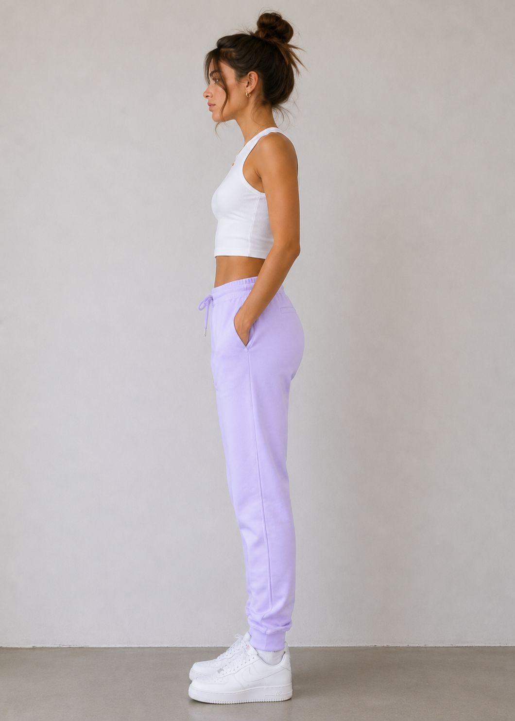 A woman in a white sports bra and lavender sweatpants stands in profile against a plain light gray wall, wearing white platform sneakers.