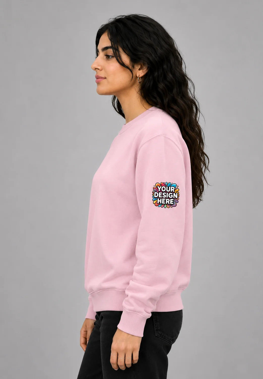 A woman in profile wearing a light pink crewneck sweatshirt with a colorful 'YOUR DESIGN HERE' mockup patch on the upper left sleeve, set against a solid gray studio background.