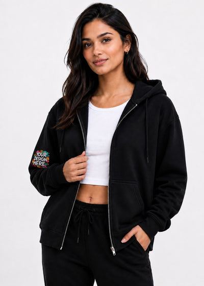 A woman modeling a black zip-up hoodie with a colorful 'YOUR DESIGN HERE!' graphic on the left sleeve, worn over a white crop top and black sweatpants against a light gray background.