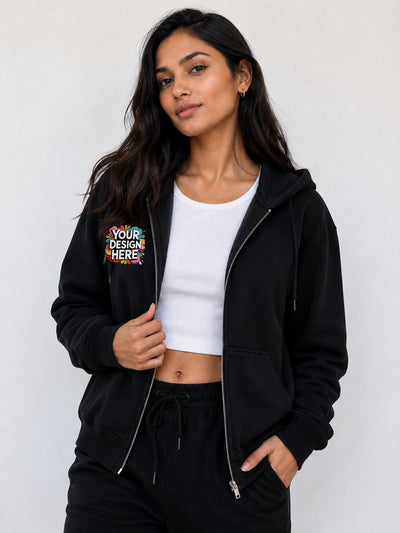 A woman modeling a black zip-up hoodie with a colorful 'YOUR DESIGN HERE' mockup graphic on the left chest, worn over a white crop top and black sweatpants against a light gray background.