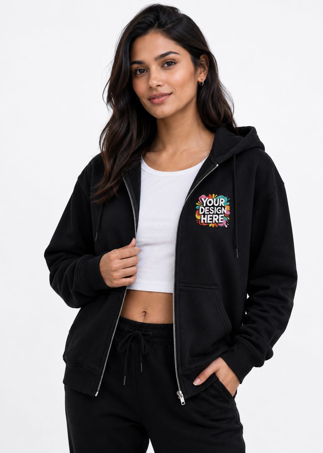 A woman modeling a black zip-up hoodie with a colorful 'YOUR DESIGN HERE' mockup graphic on the left chest, worn over a white crop top and black sweatpants against a plain white background.