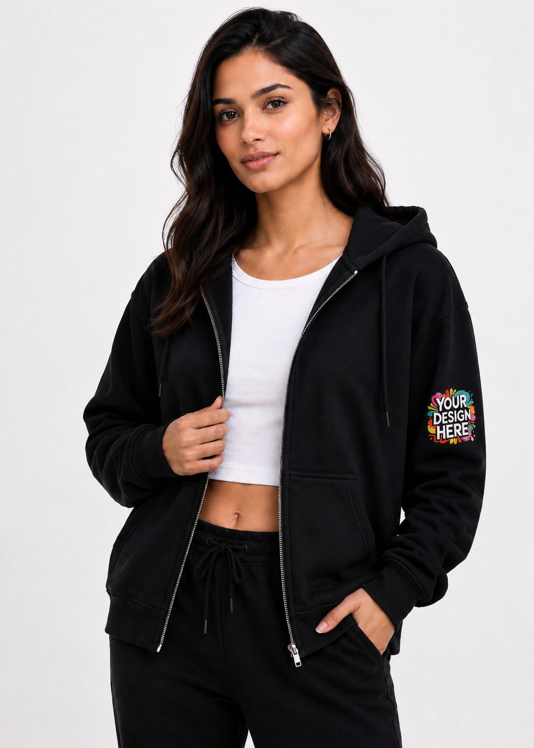 A woman modeling a black zip-up hoodie with a colorful 'YOUR DESIGN HERE!' mockup patch on the left sleeve, worn over a white crop top and black sweatpants against a light gray background.