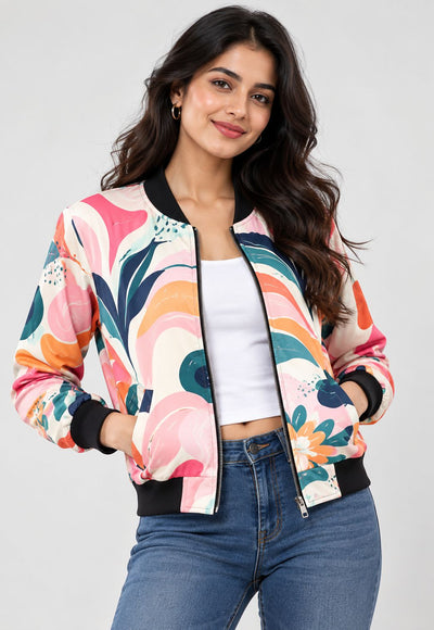 A woman modeling a colorful abstract floral bomber jacket with black ribbed trim, worn over a white crop top and blue jeans against a light gray studio background.