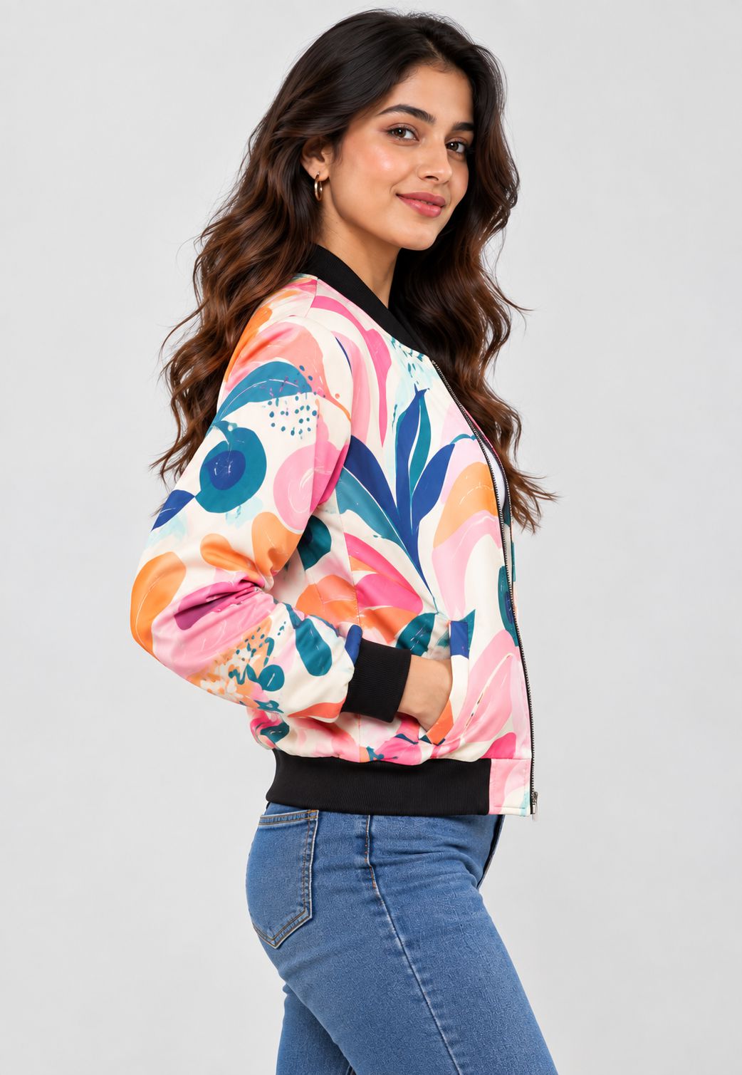 A woman modeling a colorful abstract floral bomber jacket with pink, orange, blue, and teal patterns on a white base, paired with blue jeans against a light gray studio background.