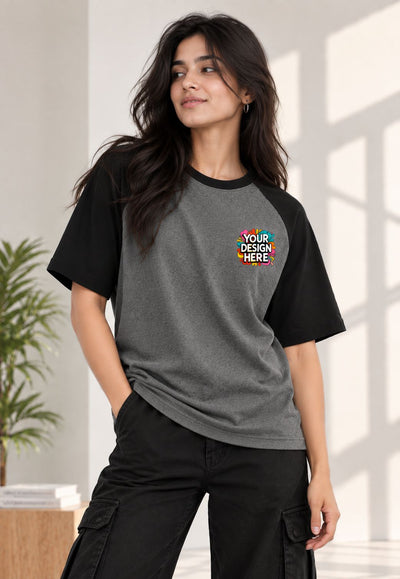 A woman modeling a custom raglan t-shirt with a dark gray body and black sleeves, featuring a colorful 'YOUR DESIGN HERE' placeholder graphic on the left chest. She is wearing black cargo pants in a bright, modern room with soft window shadows.