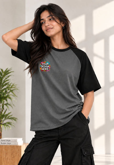 A woman modeling a dark gray and black raglan t-shirt with a colorful 'YOUR DESIGN HERE' mockup graphic on the left chest, paired with black cargo pants in a bright indoor setting.