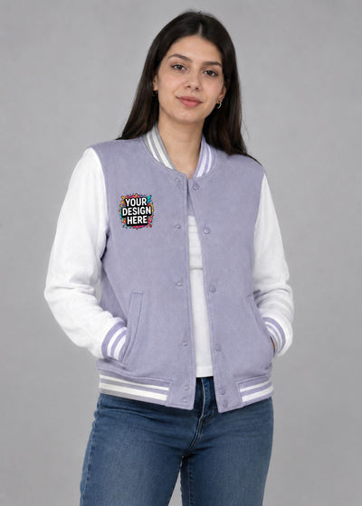 A woman modeling a lavender and white varsity jacket with a colorful 'YOUR DESIGN HERE' mockup patch on the left chest, paired with blue jeans against a solid gray studio background.