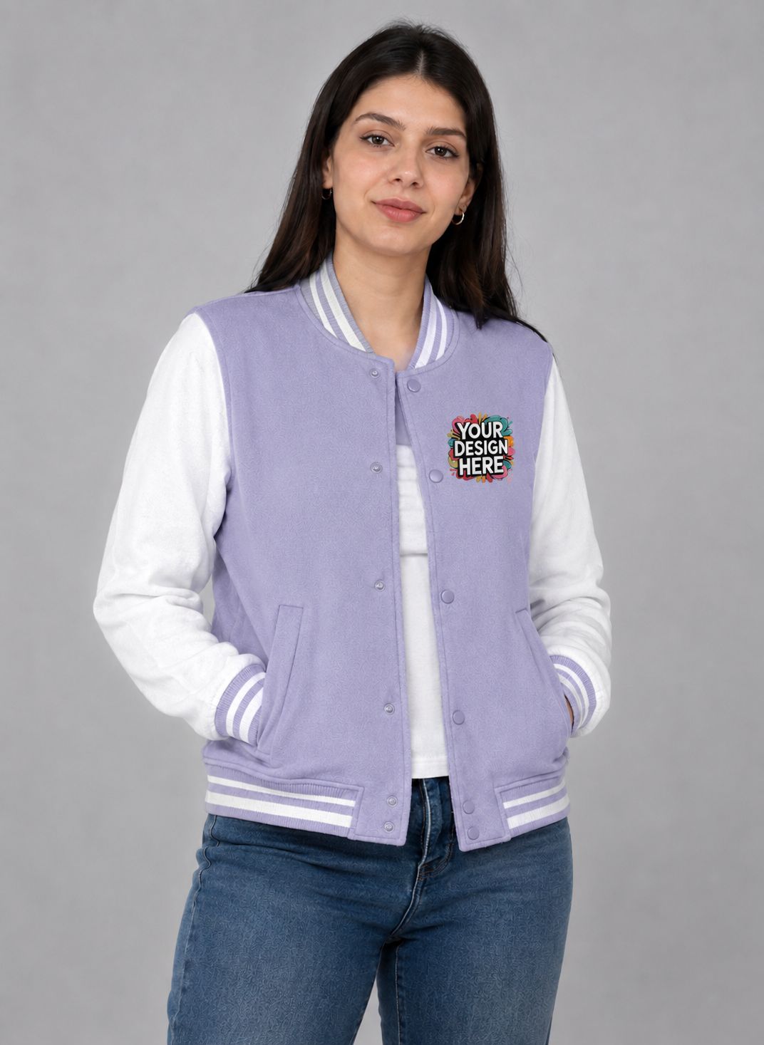 A woman modeling a lavender and white varsity jacket with a colorful 'Your Design Here' patch on the chest, paired with blue jeans against a solid gray background.
