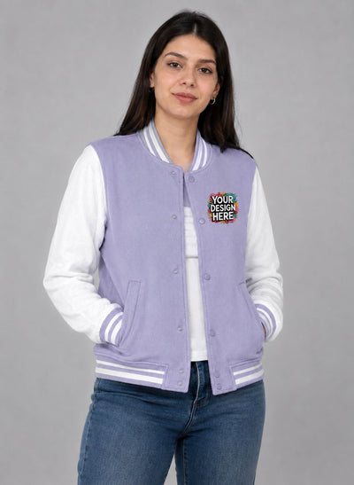 A woman modeling a lavender and white varsity jacket with a colorful 'Your Design Here' patch on the chest, paired with blue jeans against a solid gray background.