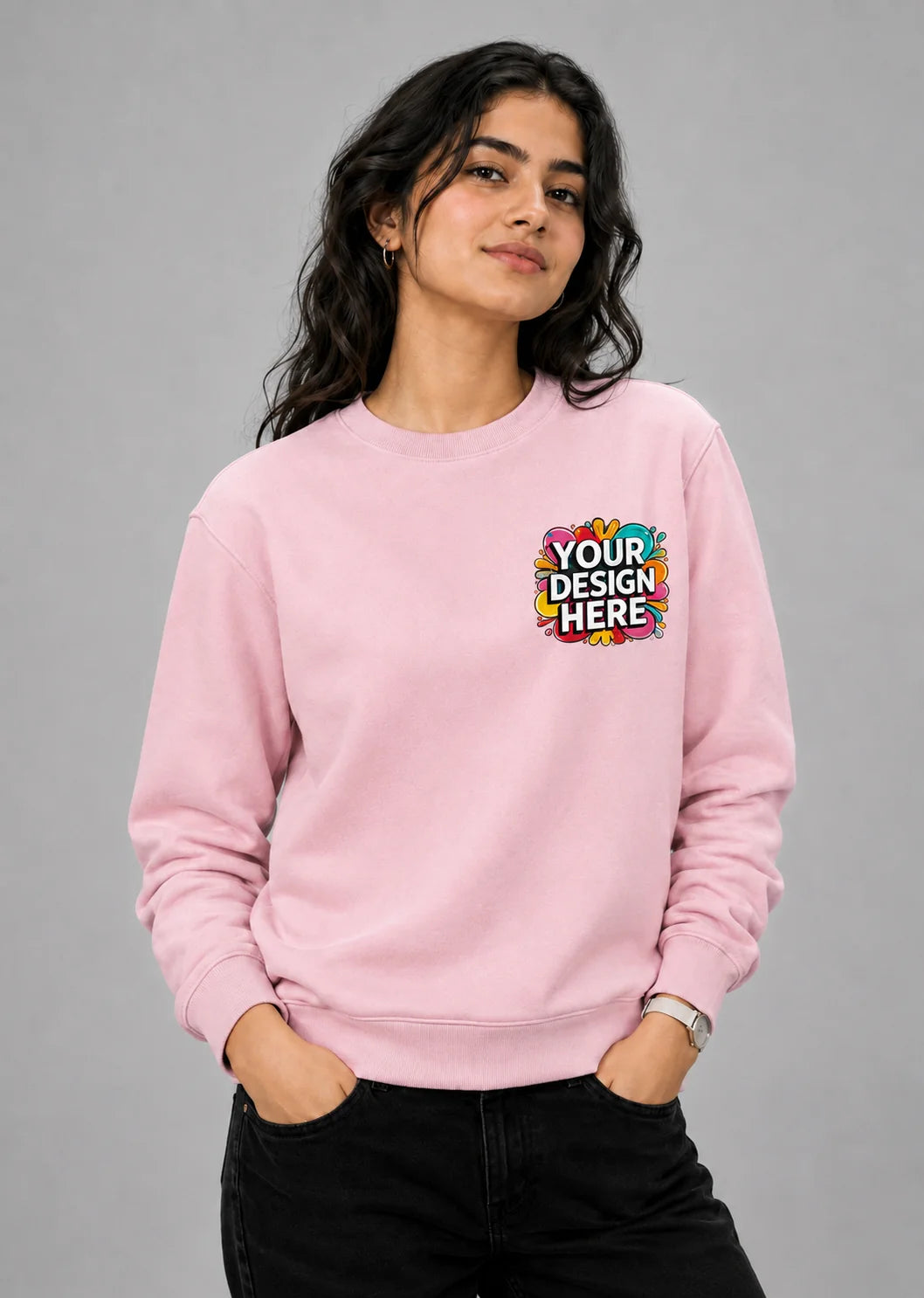 A woman modeling a light pink crewneck sweatshirt with a colorful 'YOUR DESIGN HERE' mockup graphic on the left chest, paired with black jeans against a solid gray studio background.