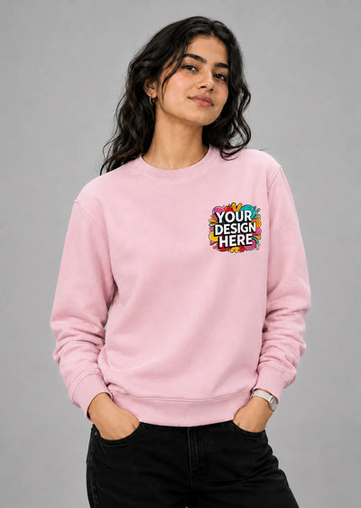 A woman modeling a light pink crewneck sweatshirt with a colorful 'YOUR DESIGN HERE' mockup graphic on the left chest, paired with black jeans against a solid gray studio background.