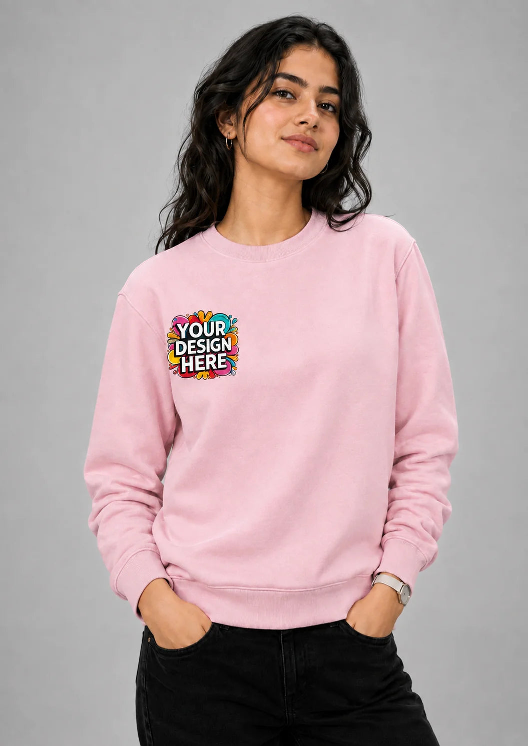 A woman modeling a light pink crewneck sweatshirt with a colorful 'YOUR DESIGN HERE' mockup graphic on the left chest, paired with black pants against a solid gray studio background.