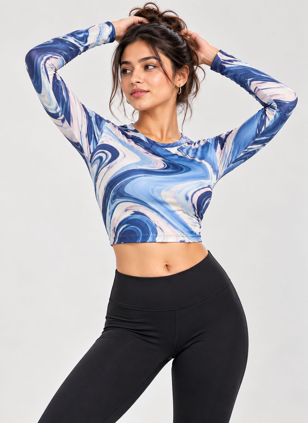 A woman modeling a long-sleeve crop top with a blue, white, and light gray marble swirl pattern, paired with black leggings, against a light gray studio background.