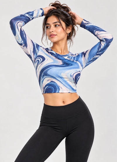A woman modeling a long-sleeve crop top with a blue, white, and light gray marble swirl pattern, paired with black leggings, against a light gray studio background.