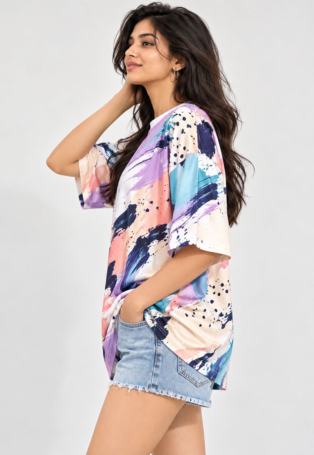 A woman modeling a loose short-sleeve t-shirt with a colorful abstract brushstroke print in purple, blue, pink, and cream, paired with denim shorts against a light gray background.