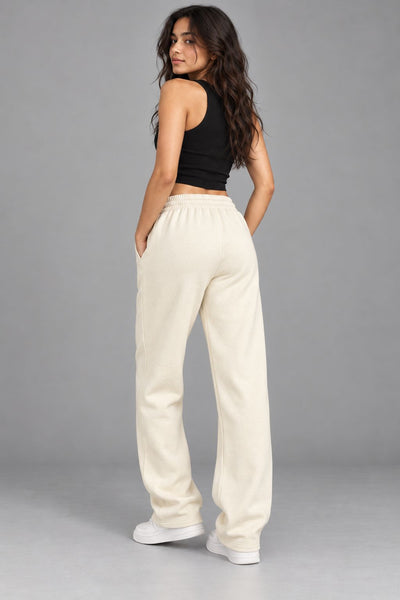 A woman modeling a pair of cream-colored, wide-leg sweatpants with a matching elastic waistband, paired with a black crop top and white sneakers, shown from a rear three-quarter angle against a solid gray studio background.