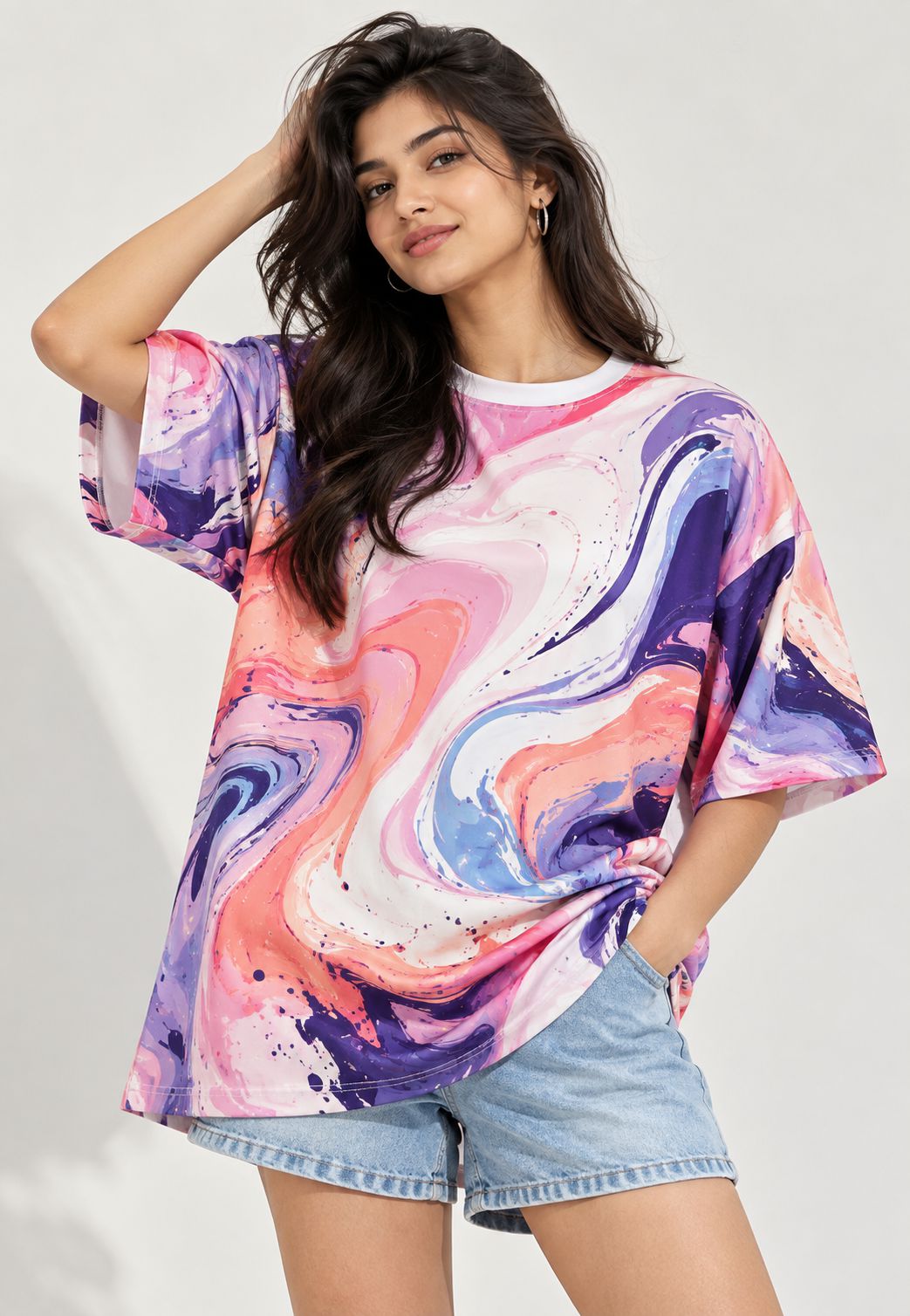 A woman modeling a short-sleeve crewneck t-shirt with a vibrant pink, purple, and blue marble swirl pattern, paired with light-wash denim shorts against a light gray background.