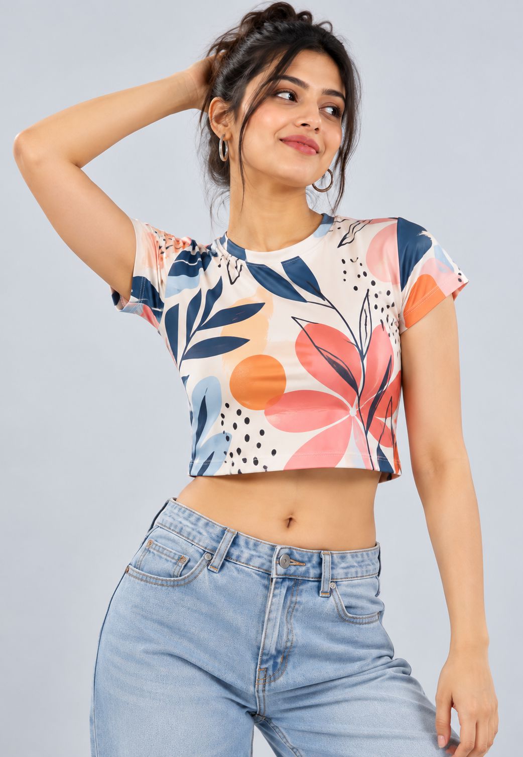 A woman modeling a short-sleeve crop top with a colorful abstract floral print in orange, pink, navy, and blue on a cream background, paired with light blue jeans against a solid gray studio background.