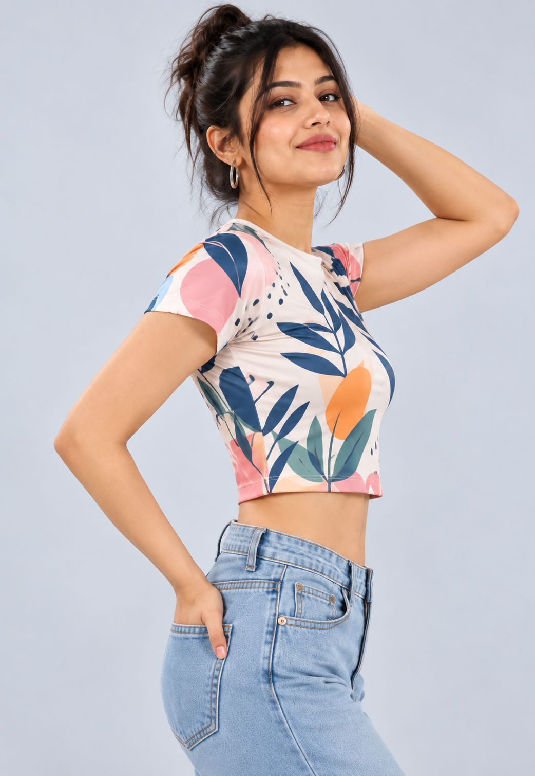 A woman modeling a short-sleeve crop top with a colorful botanical leaf pattern in pink, orange, blue, and green on a cream background, paired with light blue jeans against a solid light gray background.