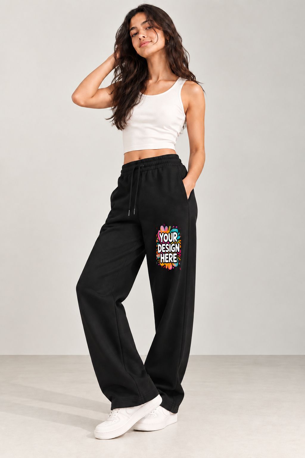 A woman modeling black wide-leg sweatpants with a colorful 'YOUR DESIGN HERE' mockup graphic on the left thigh, paired with a white crop top and white sneakers, against a light gray studio background.