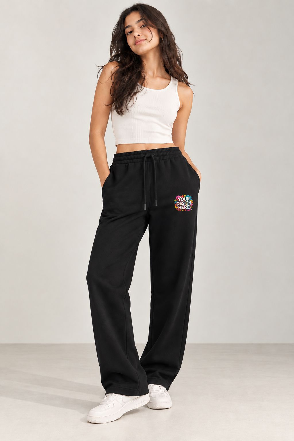 A woman modeling black wide-leg sweatpants with a colorful 'Your design here' graphic on the left thigh, paired with a white crop top and white sneakers, against a light gray studio background.