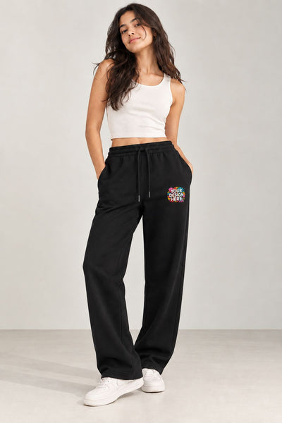 A woman modeling black wide-leg sweatpants with a colorful 'Your design here' graphic on the left thigh, paired with a white crop top and white sneakers, against a light gray studio background.