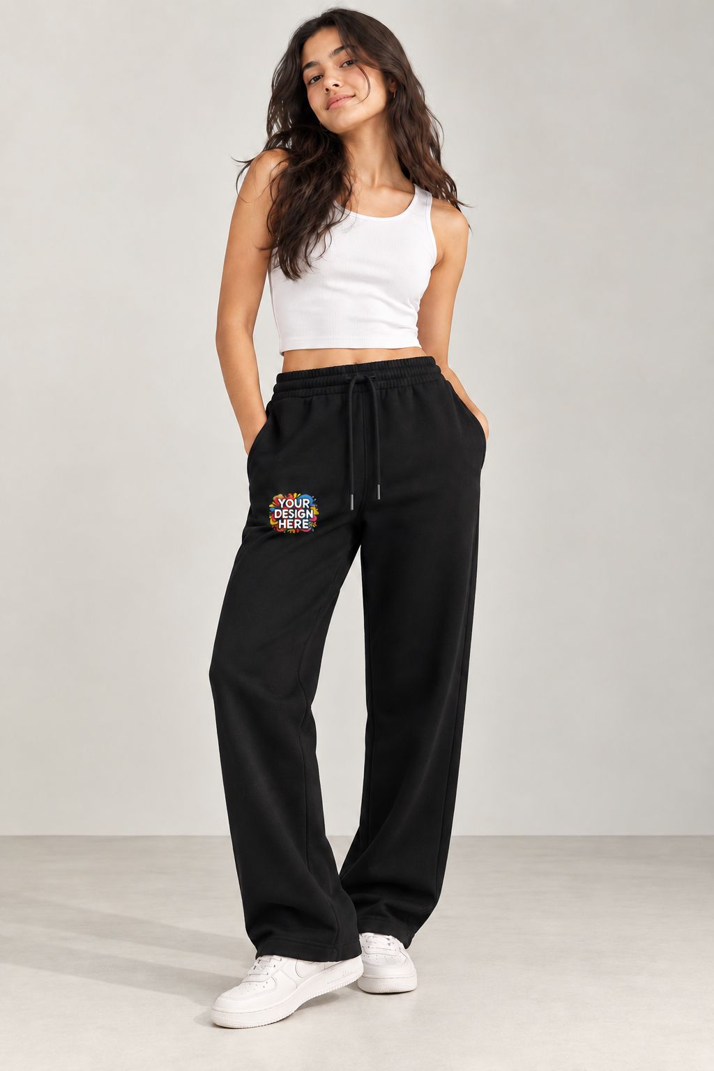A woman modeling black wide-leg sweatpants with a colorful graphic patch on the left thigh, paired with a white crop top and white sneakers, against a light gray studio background.