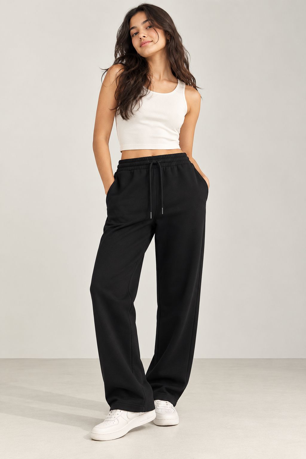 A woman modeling black wide-leg sweatpants with a drawstring waist, paired with a white crop top and white sneakers, standing against a light gray studio background.