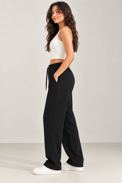 A woman modeling black wide-leg sweatpants with a drawstring waist, paired with a white crop top and white sneakers, standing against a light gray studio background.