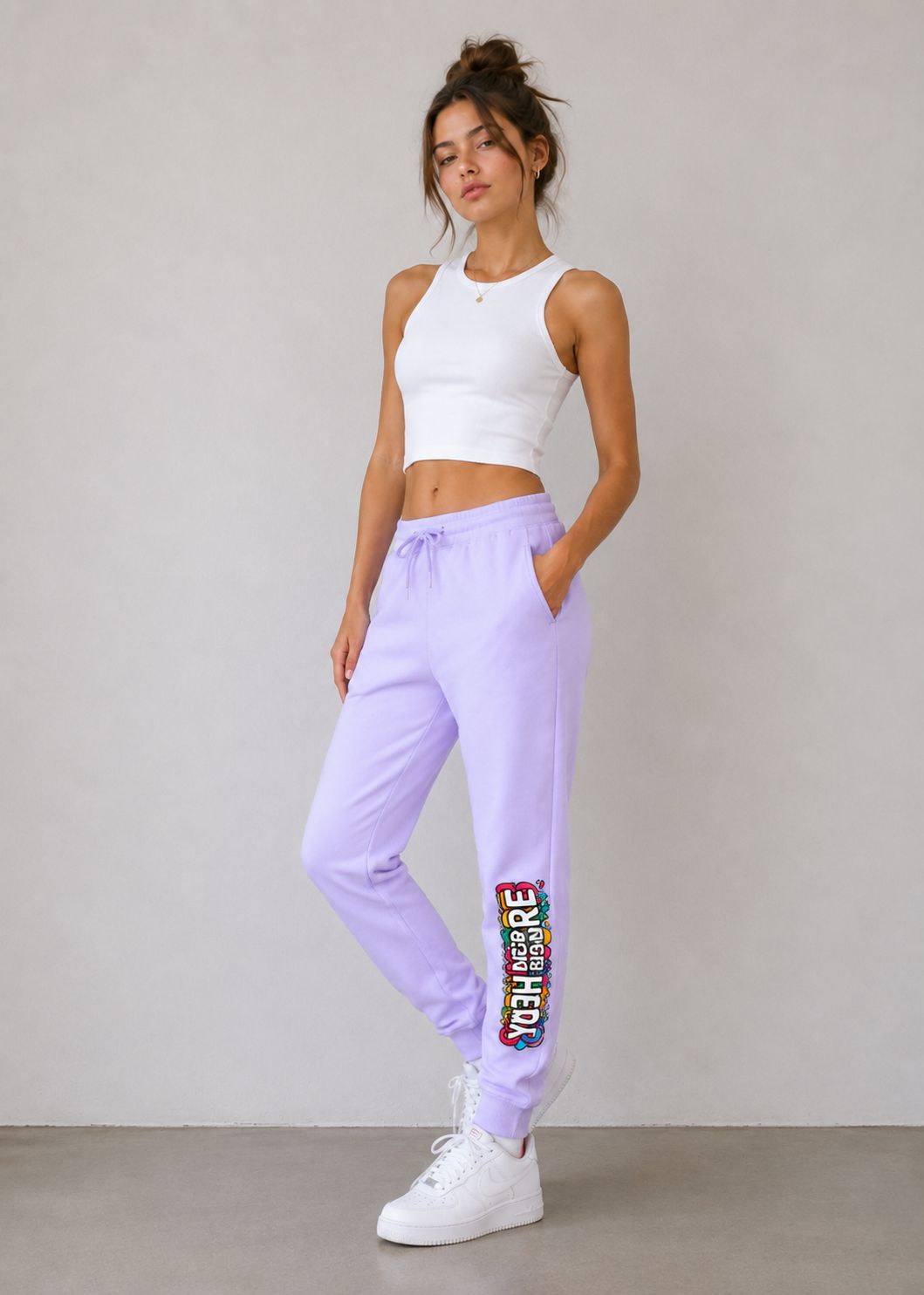 A woman modeling lavender jogger sweatpants with a colorful 'TOGETHER' graphic on the lower left leg, paired with a white crop top and white sneakers, standing against a neutral gray wall.
