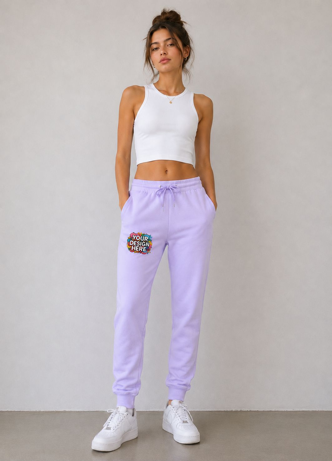 A woman modeling light purple sweatpants with a colorful 'YOUR DESIGN HERE' patch on the left thigh, paired with a white crop top and white sneakers, standing against a neutral gray wall.
