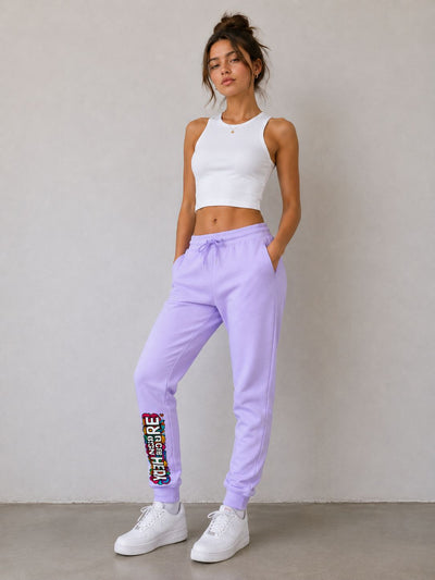 A woman modeling light purple sweatpants with a colorful graphic patch on the lower left leg, paired with a white crop top and white sneakers, standing against a neutral gray wall.