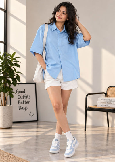 A woman models a light blue short-sleeve button-up shirt, white shorts, white socks, and light blue and white sneakers in a bright, modern room with a plant and framed art.