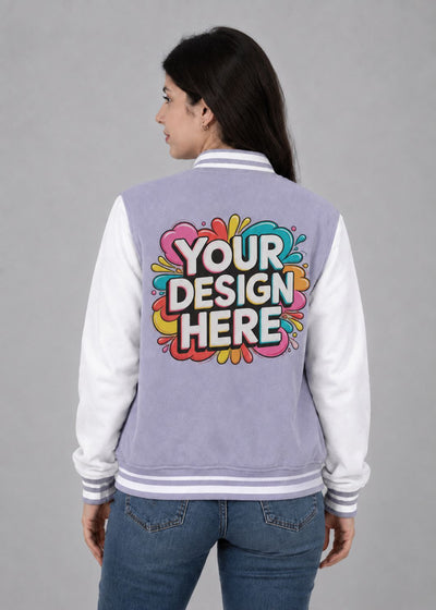 A woman shown from the back wearing a lavender and white varsity jacket with a colorful 'YOUR DESIGN HERE' graphic printed on the back, paired with blue jeans, against a solid gray studio background.