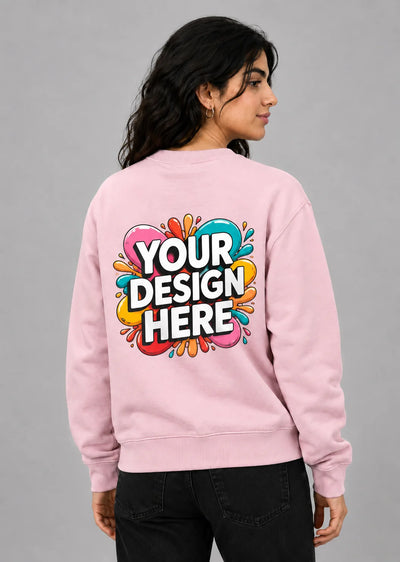 A woman shown from the back wearing a light pink crewneck sweatshirt with a colorful mockup graphic on the back that reads 'YOUR DESIGN HERE' in white letters with black outlines, set against a solid gray studio background.
