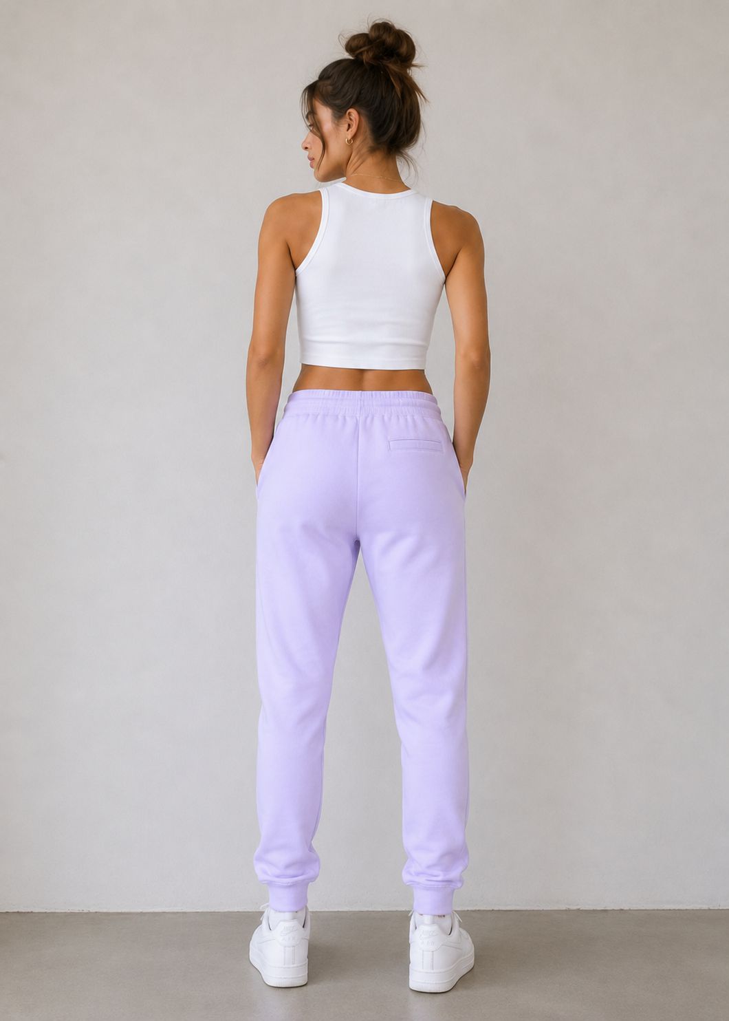 A woman shown from the back wearing light purple sweatpants and a white crop top, standing against a plain light gray wall.