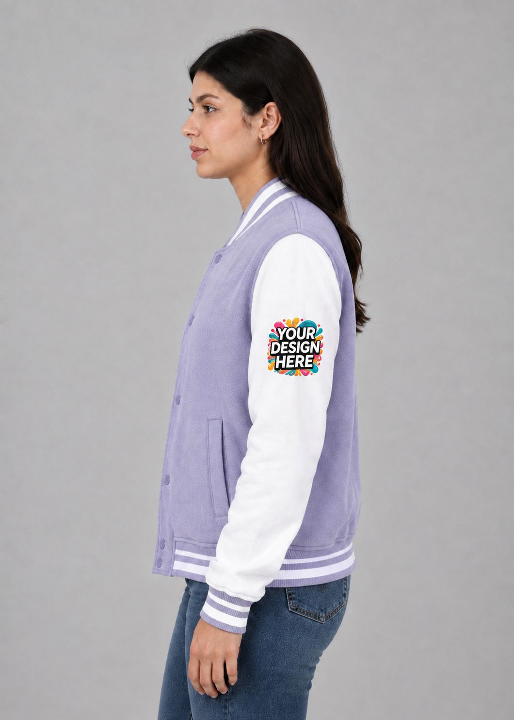 A woman shown in profile wearing a lavender and white varsity jacket with a colorful 'YOUR DESIGN HERE' mockup patch on the sleeve, paired with blue jeans, against a plain gray studio background.