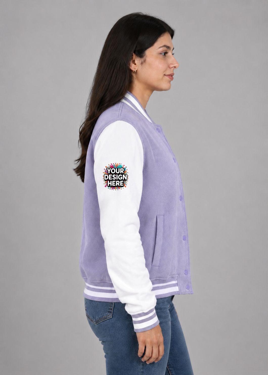 A woman shown in side profile wearing a lavender and white varsity jacket with blue jeans, photographed against a plain gray studio background. A mockup-style patch reading 'YOUR DESIGN HERE' is visible on the sleeve.