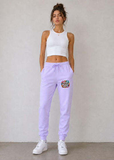 A woman stands against a light gray wall wearing lavender jogger sweatpants with a colorful 'YOUR DESIGN HERE' patch on the left thigh, a white sleeveless crop top, and white sneakers.