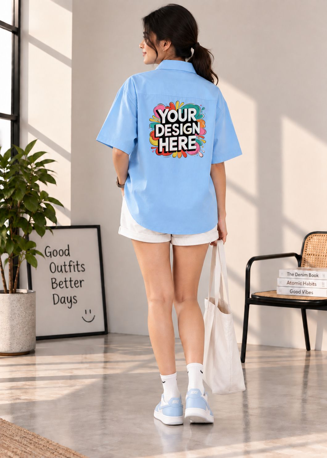 A woman stands in a bright indoor room wearing a light blue short-sleeve button-up shirt with a colorful 'YOUR DESIGN HERE' mockup graphic on the back, paired with white shorts, socks, and sneakers.