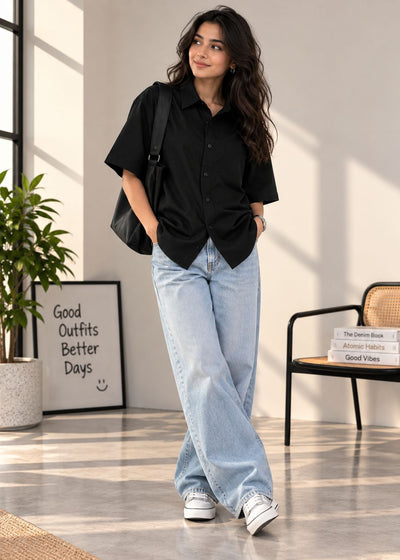 A woman stands in a bright indoor room wearing a loose black short-sleeve button-up shirt, light blue wide-leg jeans, white sneakers, and carrying a black shoulder bag.