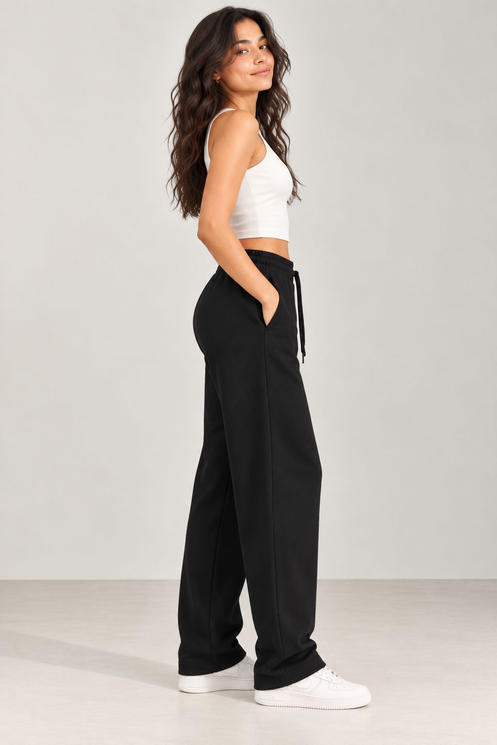 A woman stands in profile wearing black wide-leg sweatpants with a drawstring waist, a white cropped tank top, and white sneakers against a light gray studio background.
