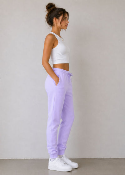 A woman stands in profile wearing light lavender sweatpants, a white crop top, and white sneakers against a plain light gray wall.