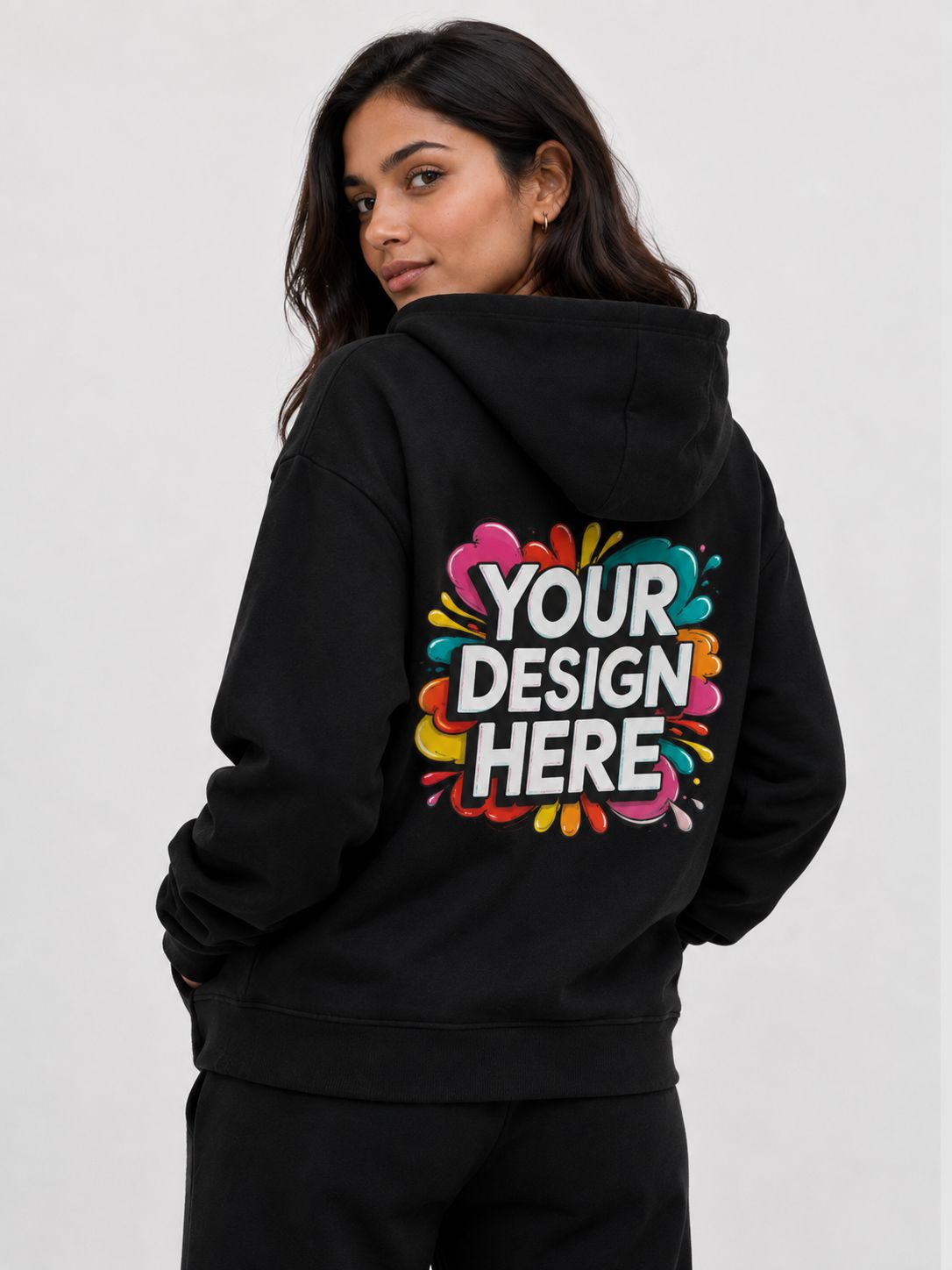A woman wearing a black hoodie from the back, featuring a colorful paint-splatter graphic with the text 'YOUR DESIGN HERE' printed on the back. The background is a clean, light gray studio backdrop.