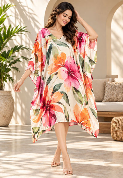 A woman wearing a flowing kaftan dress with a vibrant tropical floral print in pink, orange, and green on a cream background, walking in a sunlit indoor patio setting.