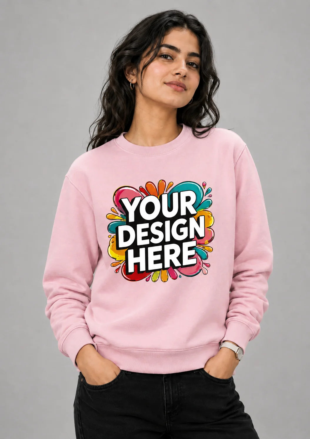A woman wearing a light pink crewneck sweatshirt with a colorful 'YOUR DESIGN HERE' mockup graphic on the front, paired with black pants, against a solid gray studio background.