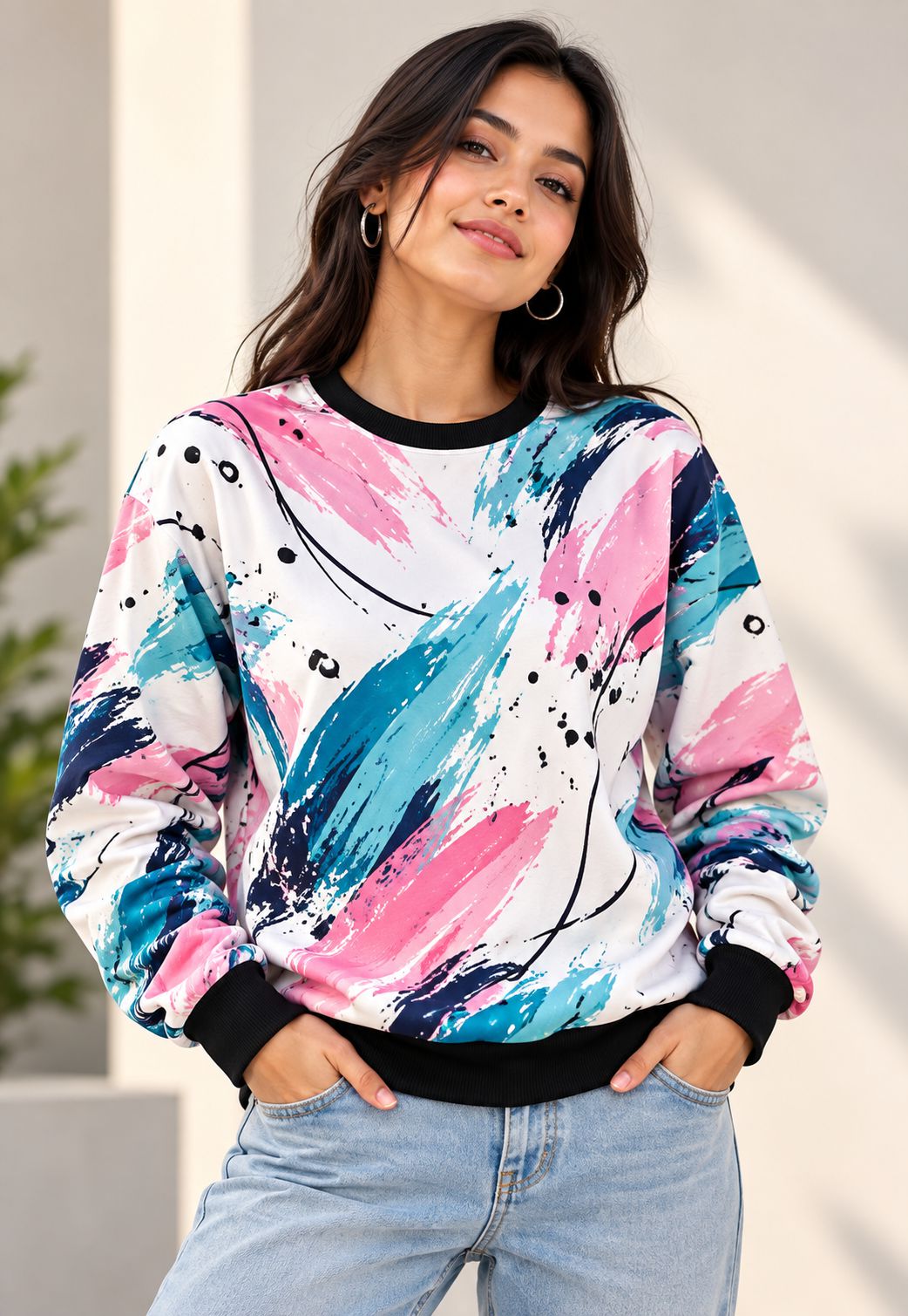 A woman wearing a long-sleeve crewneck sweatshirt with a colorful abstract paint stroke pattern in pink, blue, and teal on a white background, paired with light blue jeans.