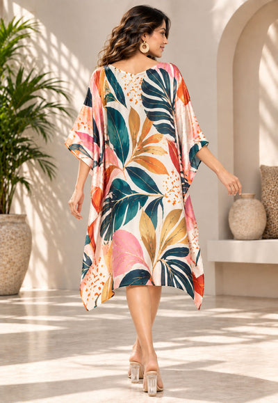 A woman wearing a loose, knee-length kaftan dress with a colorful tropical leaf print, walking in a sunlit indoor courtyard with plants and arched walls.