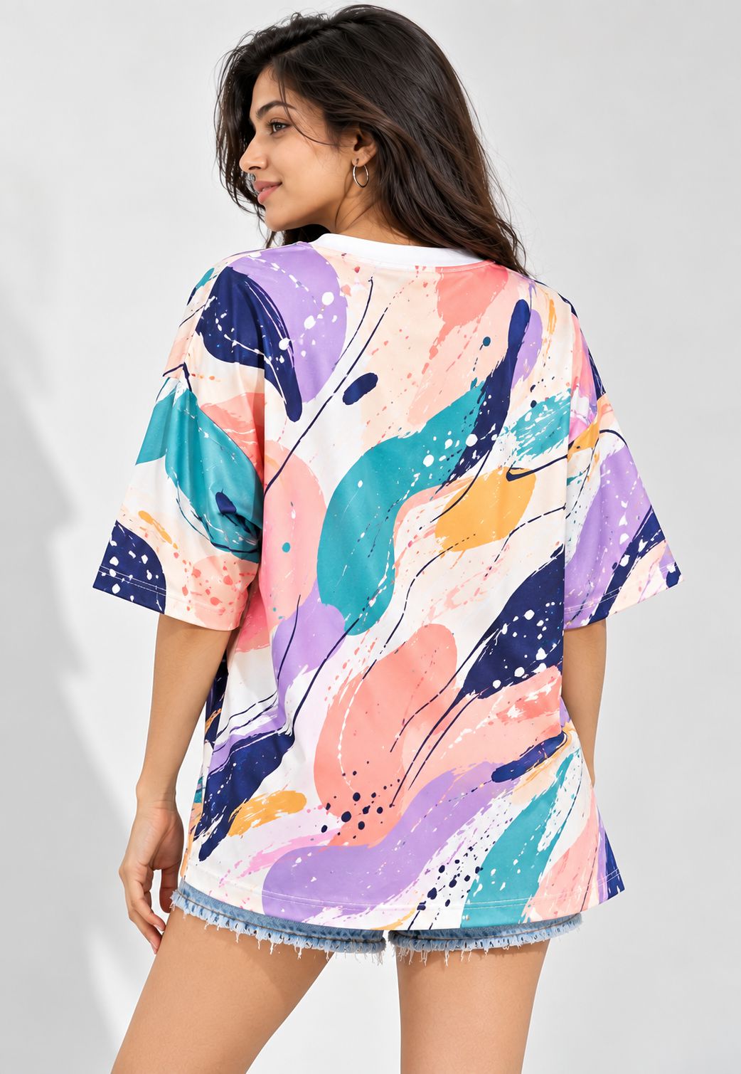 A woman wearing a loose short-sleeve t-shirt with a colorful abstract paint splatter pattern in purple, teal, pink, and orange, paired with denim shorts, shown from a three-quarters back view against a light gray background.