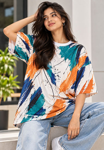 A woman wearing a loose white t-shirt with a colorful abstract brushstroke pattern in orange, teal, and navy blue, paired with light-wash jeans, sitting on a stone ledge.